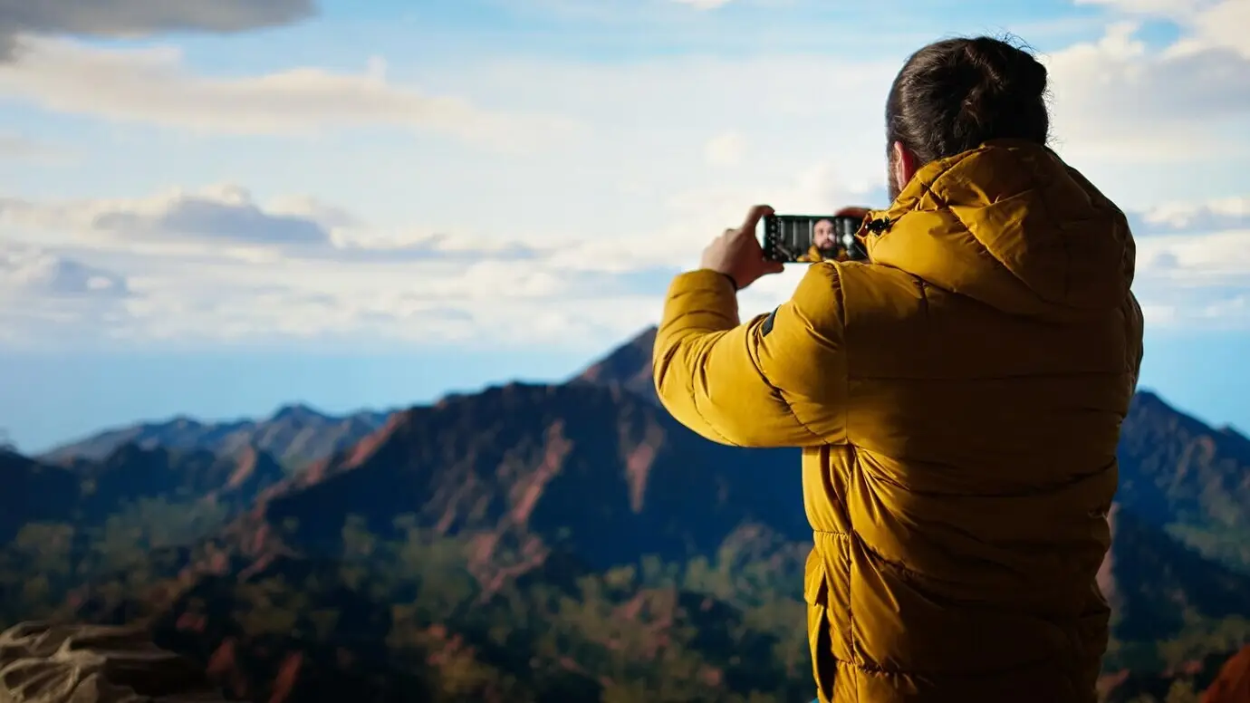 Junger abenteuerlustiger Mann, der auf einem Berggipfel mit seinem Handy Fotos macht.
